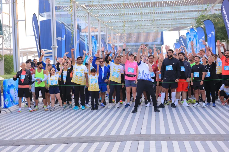 A group of participants in athletic gear, raising their hands enthusiastically at a running event, surrounded by banners and a festive atmosphere.