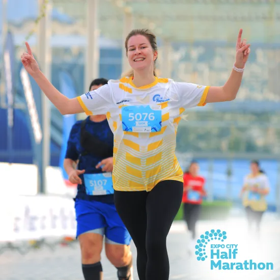 A runner in a yellow and white shirt raises both arms in celebration during the Expo City Half Marathon, with other participants in the background.