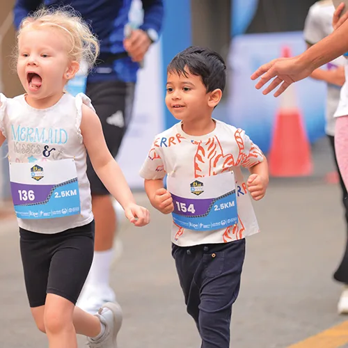 Two children, one in a white shirt and the other in a patterned tee, joyfully run during a race event.
