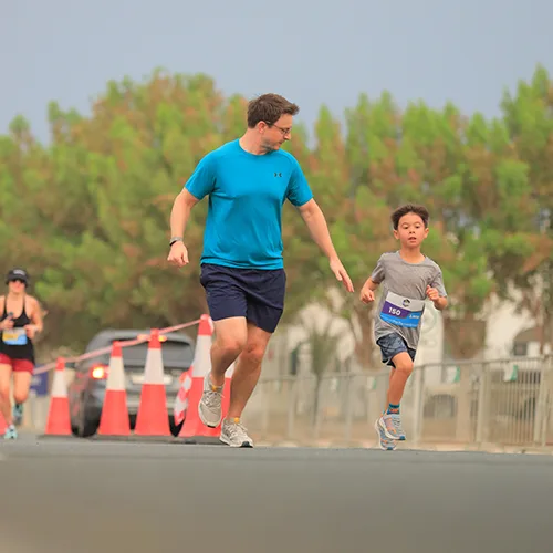 A man in a blue shirt runs alongside a young boy in a gray shirt during a race, with trees and spectators in the background.