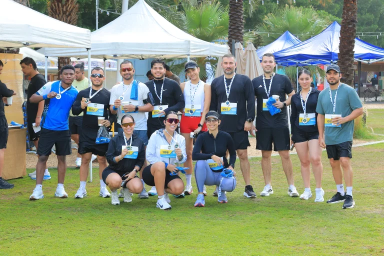 A group of participants poses with medals and bibs after a race, surrounded by tents and palm trees on a grassy field.