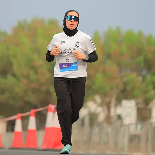A person runs in athletic gear, wearing a white shirt and dark pants, with a race number displayed, against a blurred background of trees and cones.
