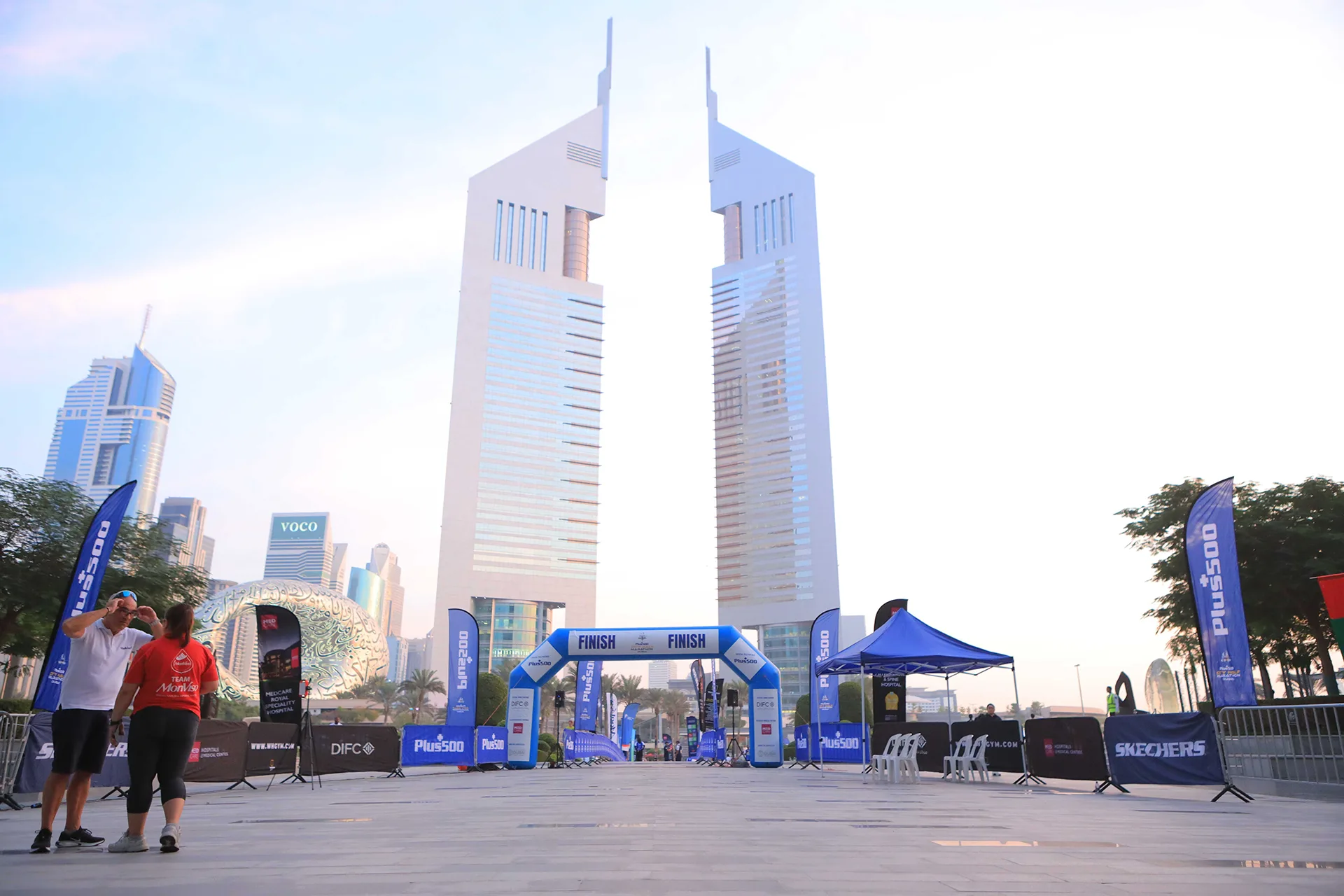 A tranquil urban scene at sunrise featuring two tall modern towers, a finish line arch, and promotional banners in a spacious plaza.