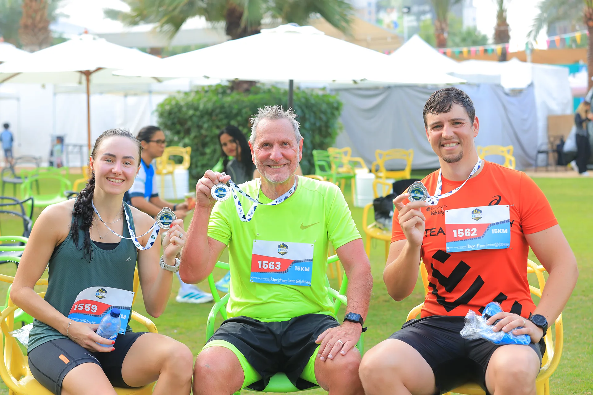Three athletes proudly display their medals after completing a race, seated outdoors amidst a festive atmosphere with palm trees.