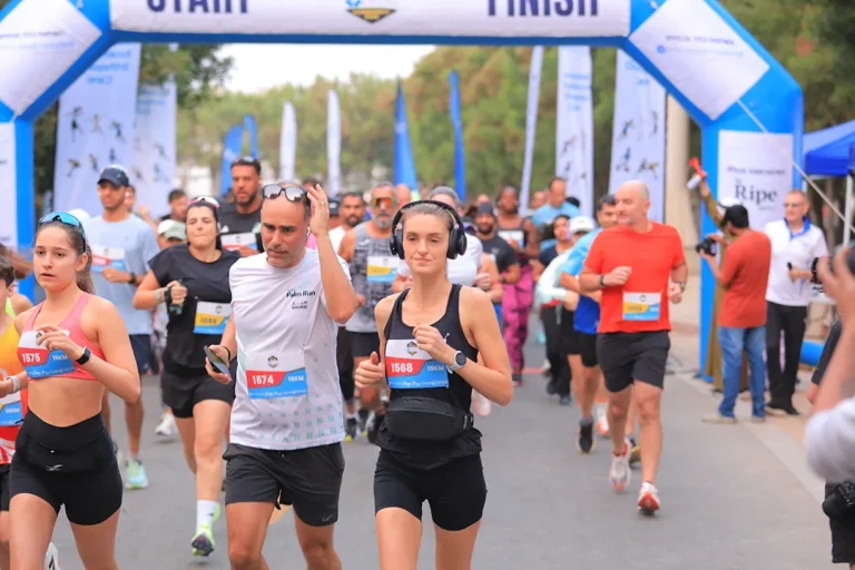 A lively group of runners at a marathon start line, wearing race numbers, with an arch marking the finish in the background.