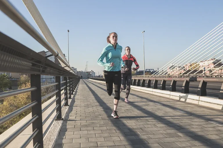 Two runners in athletic gear jog along a modern urban bridge, with clear skies and urban scenery in the background.