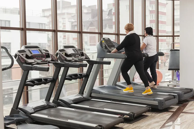 Two women using treadmills in a modern gym with large windows, showcasing a bright and spacious environment.