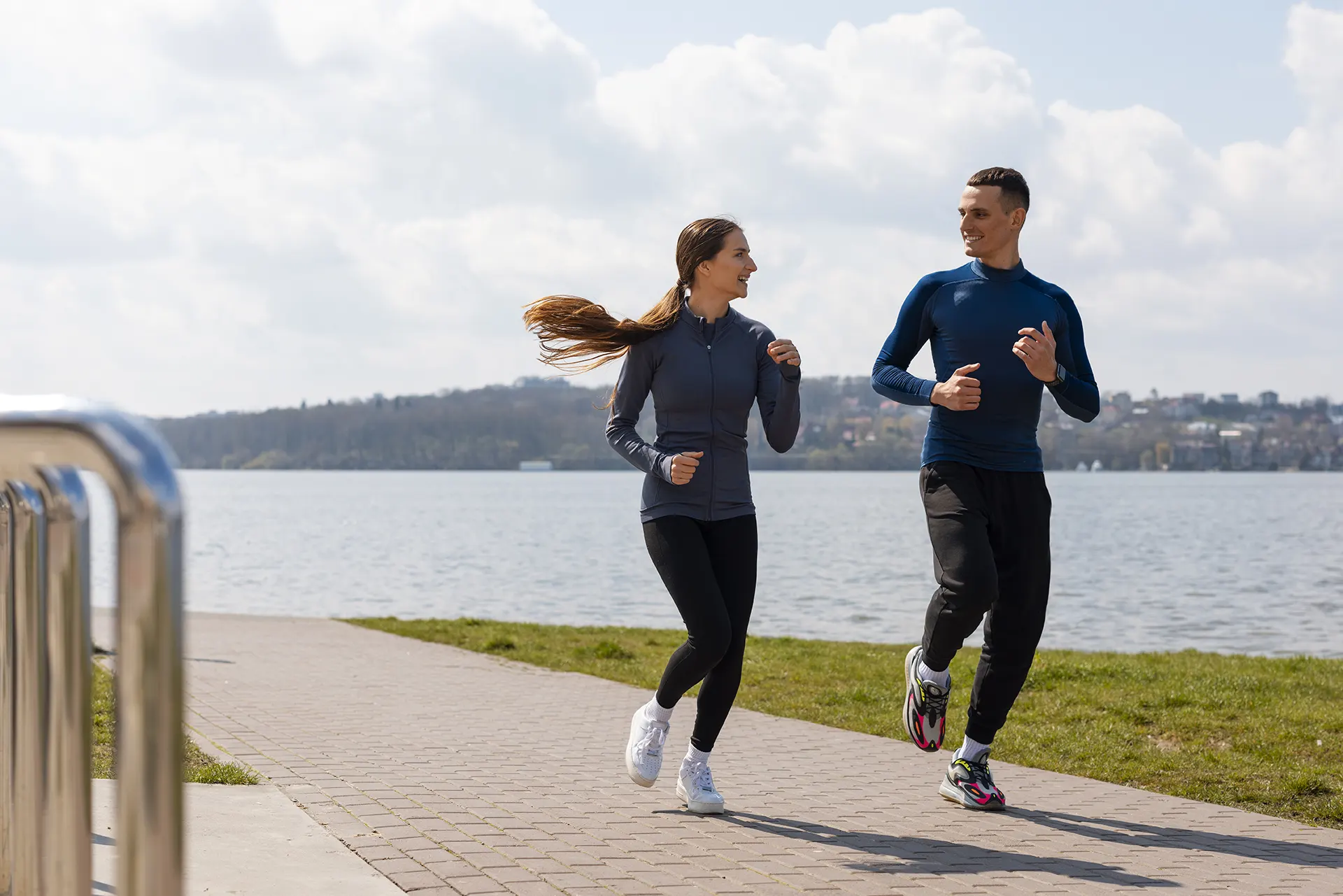 A man and woman jog along a lakeside path, with green grass and gentle waves visible under a cloudy sky.