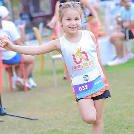 A child in athletic wear participates in a race, smiling and holding hands with another person, surrounded by spectators and chairs.