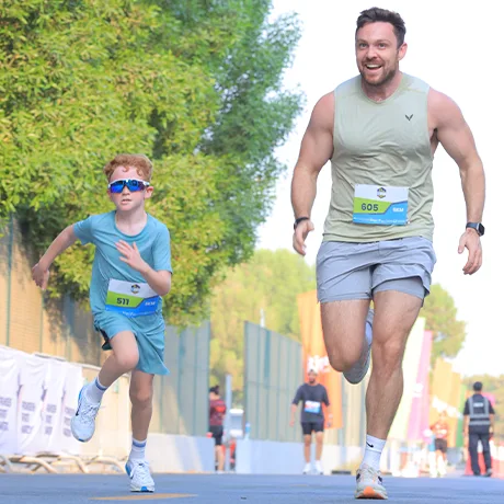 A child in a blue outfit races ahead of an adult man in a tan tank top during a running event, with greenery in the background.