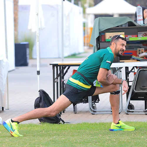 An athlete stretches on a field, wearing a green shirt, black shorts, and bright yellow sneakers, preparing for an event.