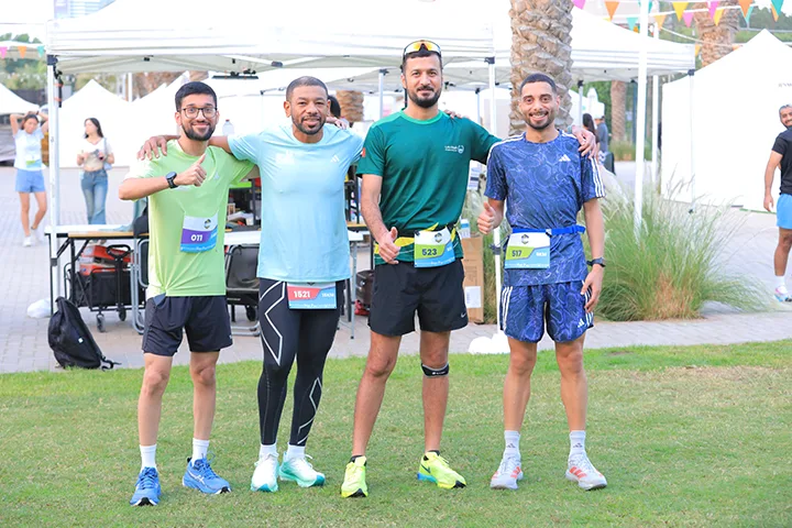 Four runners pose together, each wearing race bibs and athletic gear, with a festive atmosphere and tents in the background.