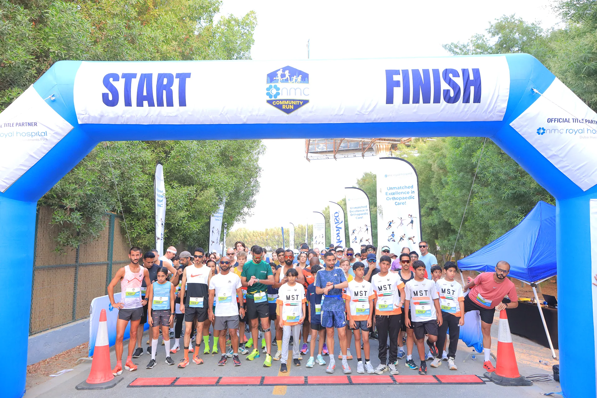 Runners gather at a start/finish line for a community race, with banners and a festive atmosphere in a green setting.