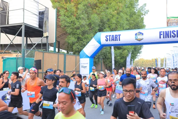 A diverse group of runners participates in a race, passing under a large "START" archway surrounded by greenery and cheering spectators.