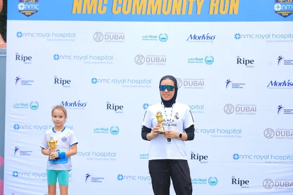 Two participants stand on a podium holding trophies at the NMC Community Run, with a promotional backdrop.