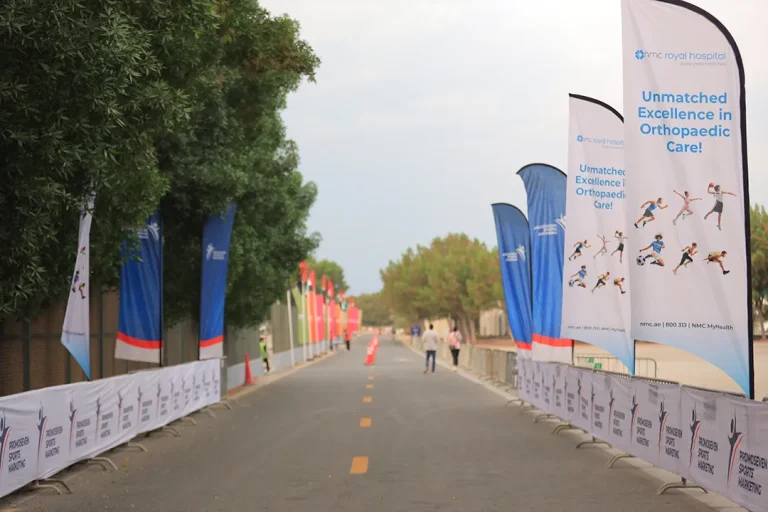 A wide path lined with banners advertising orthopaedic care, surrounded by trees, leading to a grassy area in the distance.