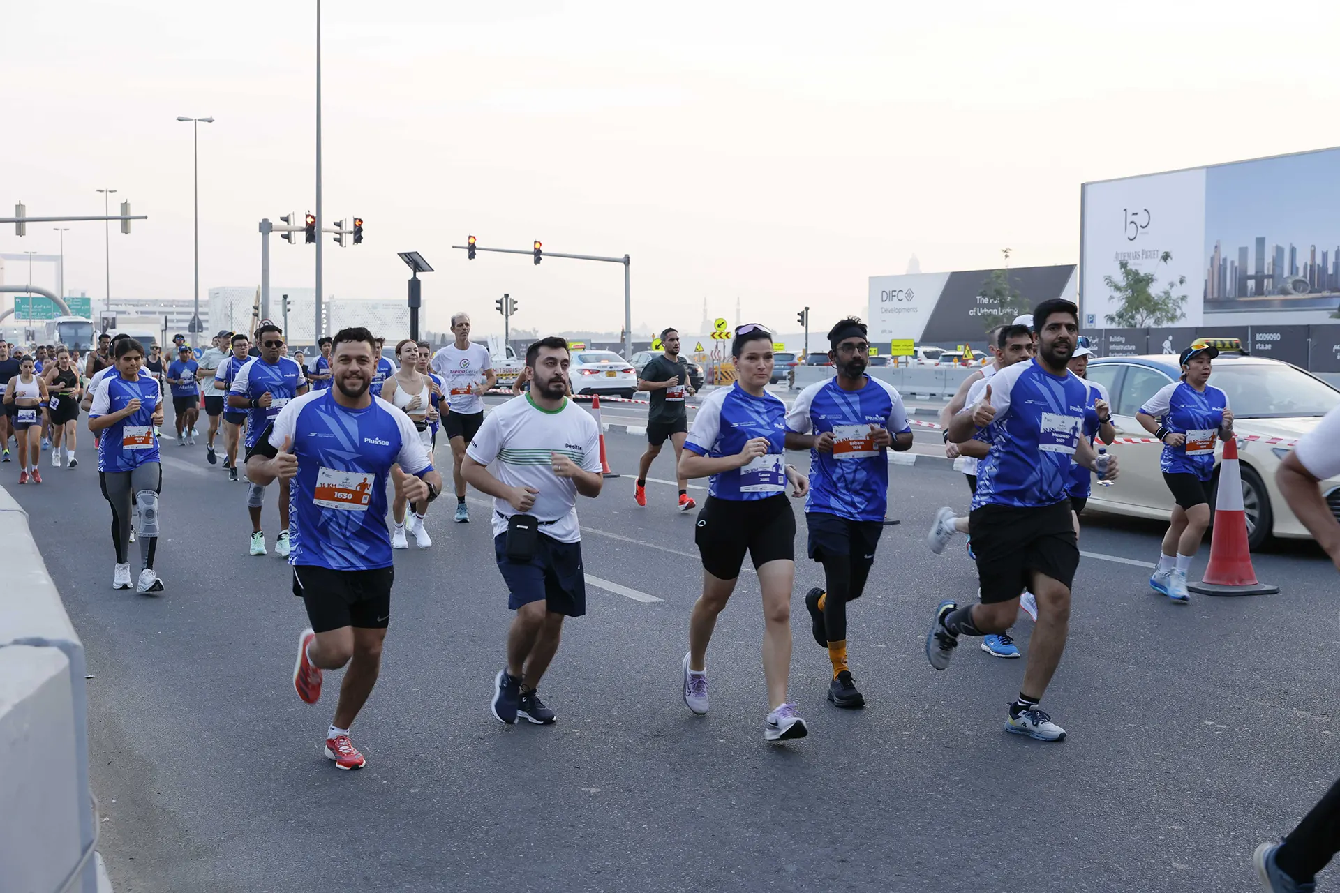 A group of runners in blue shirts participates in a road race, while cars and traffic signals line the background.