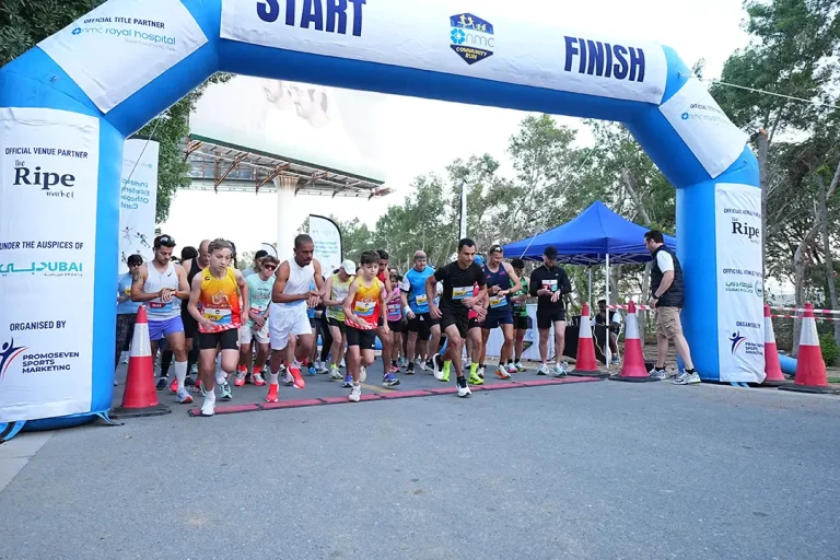 Runners start a community race, passing under a large inflatable arch marked START, with spectators and tents in the background.