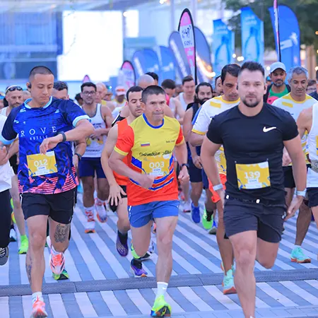 A group of runners in bright attire jogs energetically along a striped path during a race, with banners and spectators in the background.