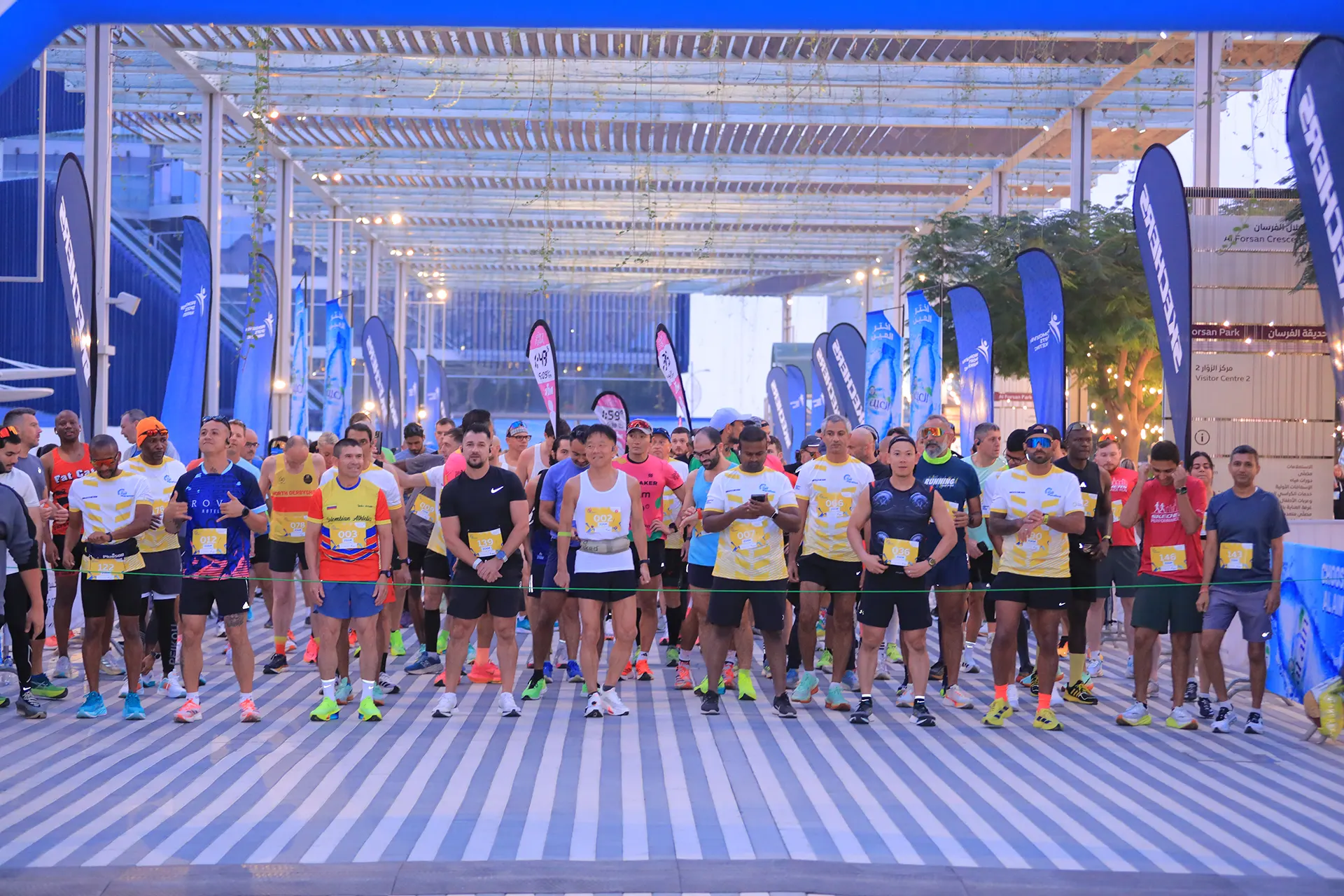 A group of runners stands at the starting line, ready for a race, surrounded by flags and a vibrant urban setting.