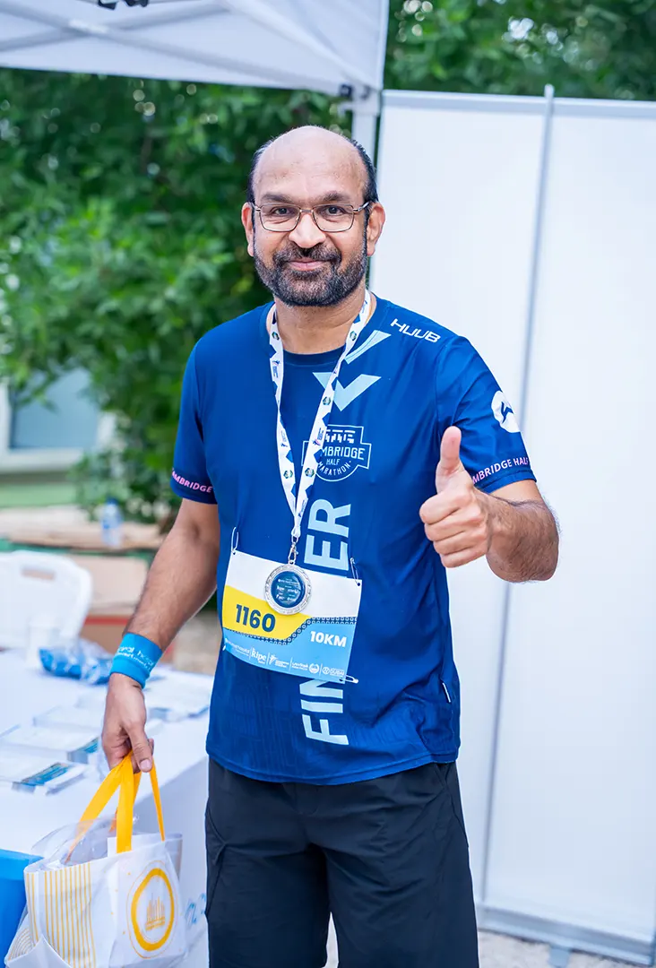 A person in a blue athletic shirt gives a thumbs up, wearing a finisher medal and holding a bag at a race event.