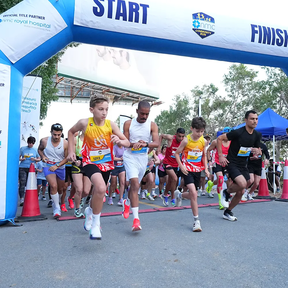 Runners in colorful attire sprinting past a blue start/finish arch during a community run event, with spectators in the background.