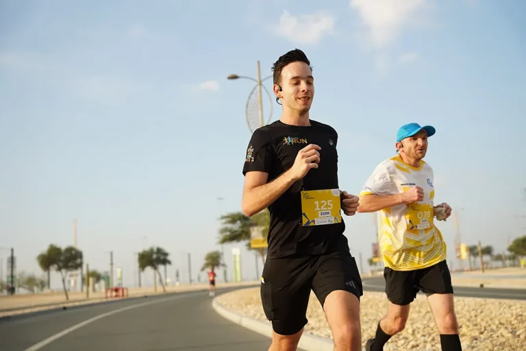 Two runners compete in a race on a sunny day, wearing numbered bibs and athletic attire, with a winding road and trees in the background.