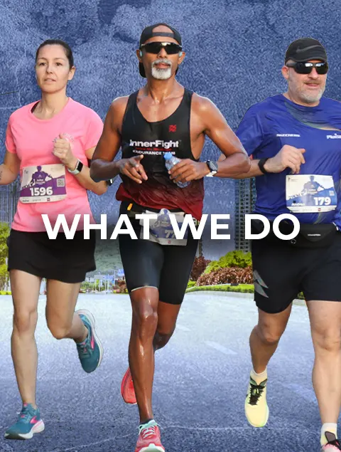 Three runners wearing race bibs running on a road, with the text WHAT WE DO overlaid in the center.