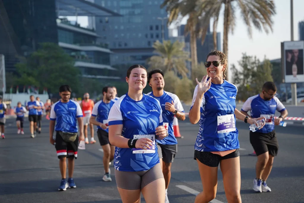Several runners wearing blue event shirts participate in a road race, with one runner waving and smiling at the camera.
