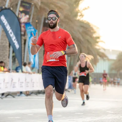 A runner in a red shirt and navy shorts participates in a race, flashing a peace sign while others run in the background.