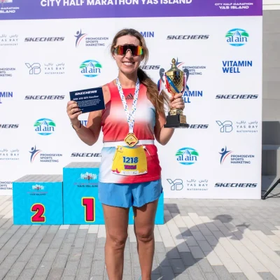 A runner proudly displays a trophy and a gift voucher, wearing a medal and sportswear, in front of a marathon backdrop with podiums.