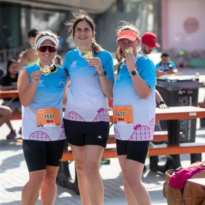 Three runners display their medals in matching blue athletic shirts, celebrating their achievement at a busy outdoor event.