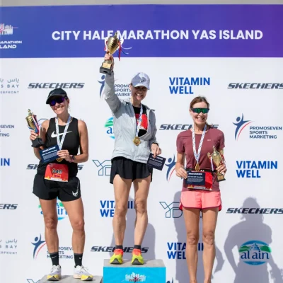 Three female marathon winners stand on podiums, holding trophies and certificates, celebrating their achievements at the Yas Island Half Marathon.