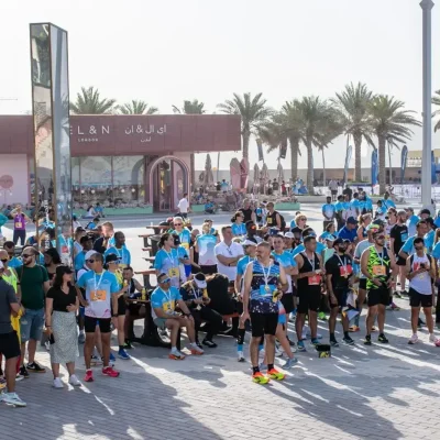 A large crowd of participants in blue shirts gathers outdoors at a lively event, with palm trees and tents in the background.